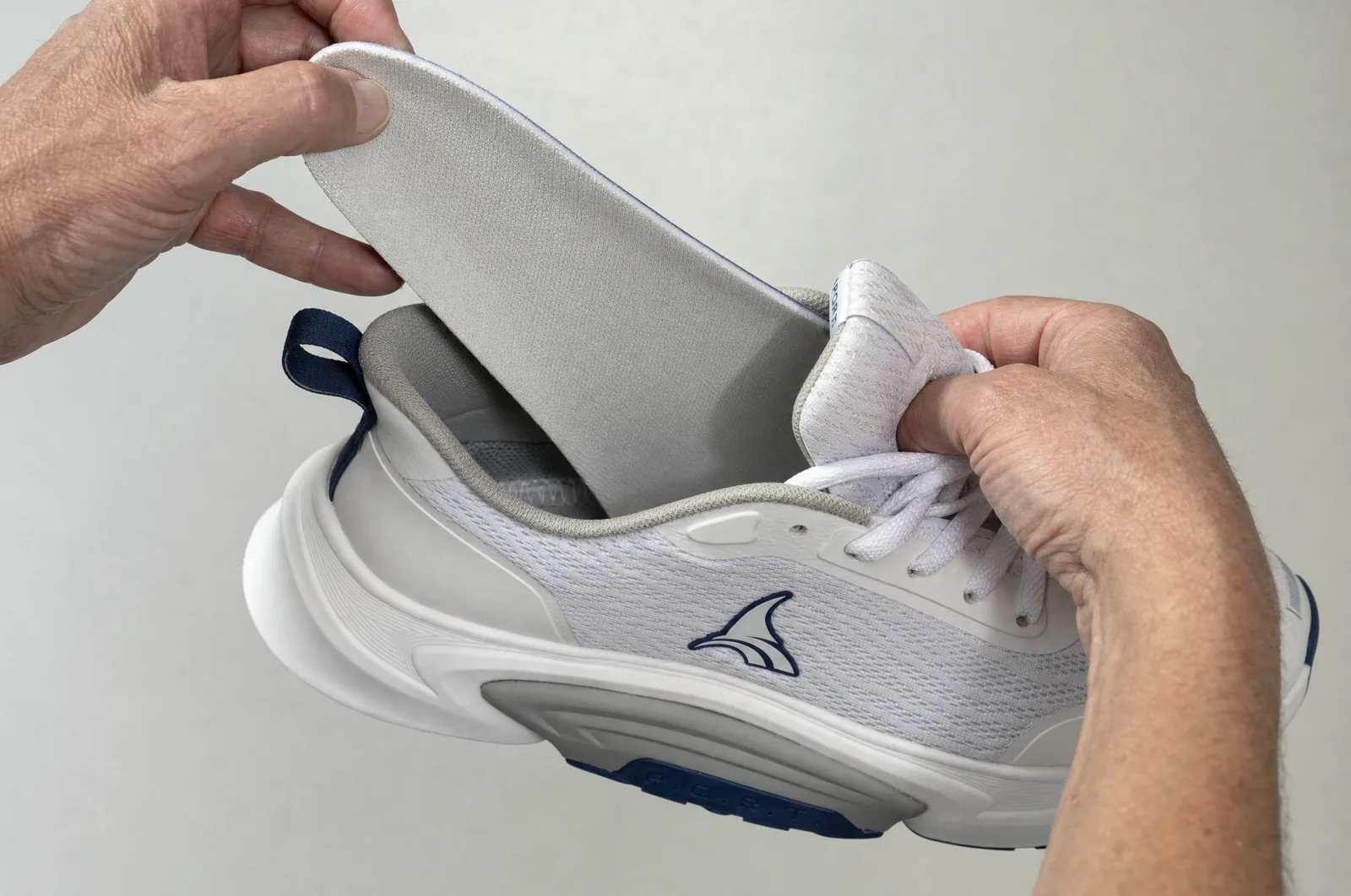The insole lifted clear of the shoe, held in one hand, the empty footbed visible below it. Product and hands only. Half sizes run to 11.5 only. No orthotic brand may appear.